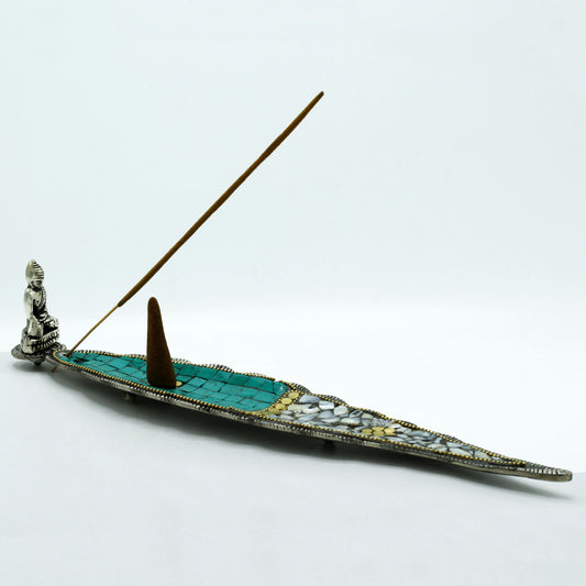 A decorative incense holder with a small figure at one end supports a lit incense stick and a cone, set against a plain white background.