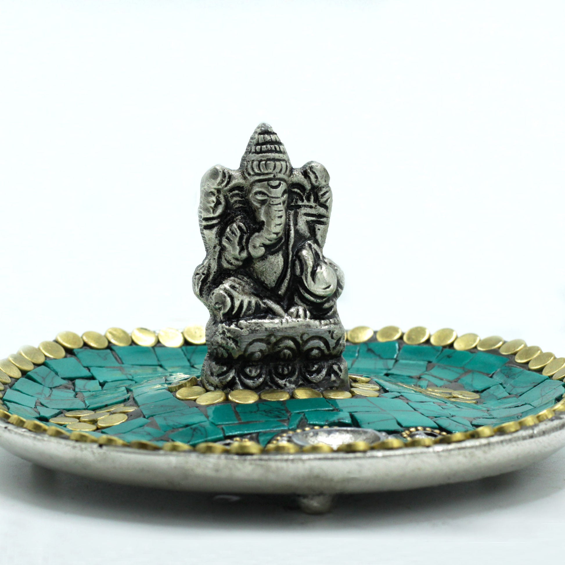 A small metal Ganesha statue sits atop a decorative plate with turquoise and gold accents, set against a plain white background.