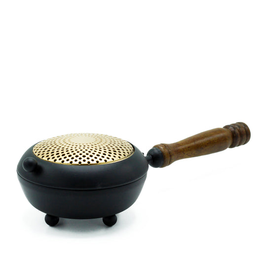 A black metal brazier with a perforated brass lid rests on small legs, featuring a wooden handle. It is placed on a white background.