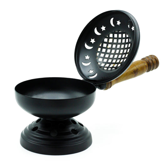 A black incense burner with a round bowl and star-moon-patterned perforated lid, featuring a wooden handle, sits in a bright, minimalistic setting.
