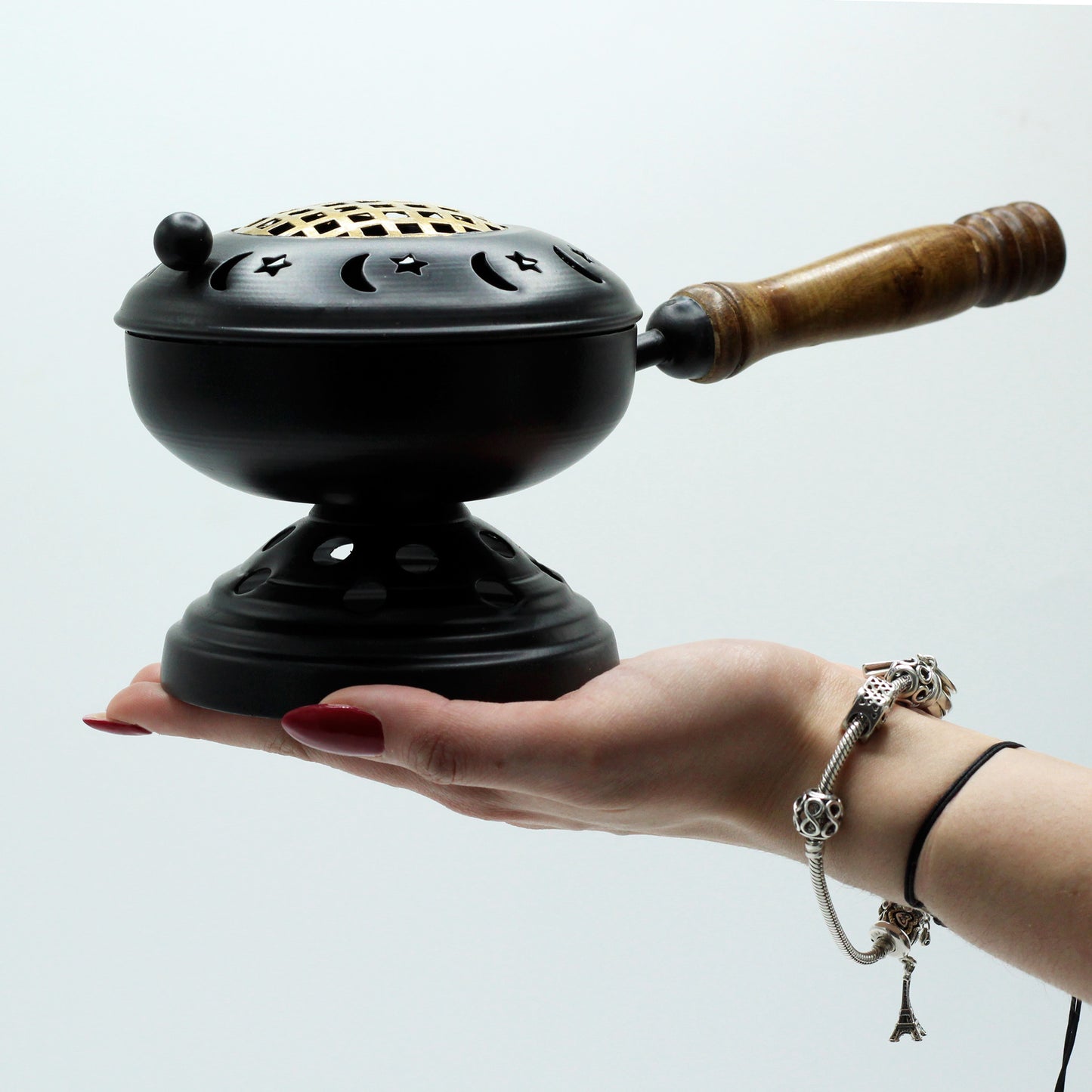 A black incense burner with star and moon cutouts rests on an outstretched hand. It features a wooden handle and is set against a plain white background.