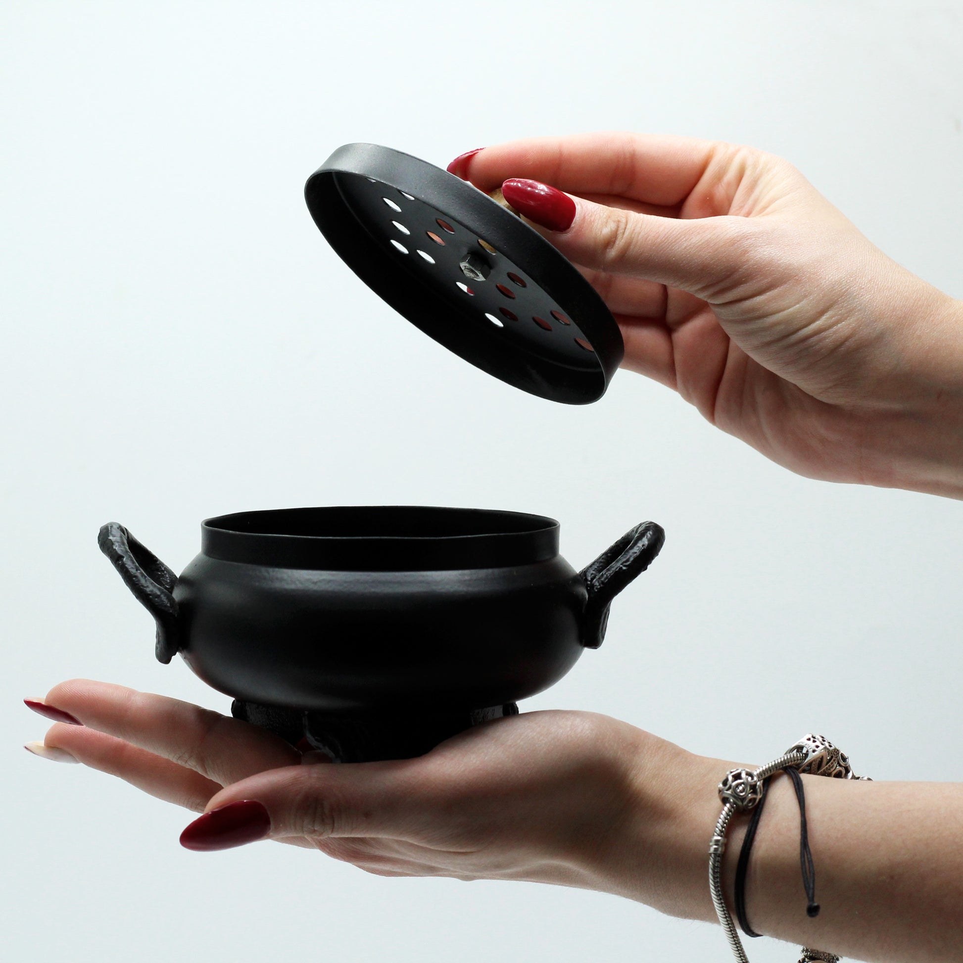 A black cauldron with two side handles is held in one hand, while the other hand removes its perforated lid. The background is plain white.