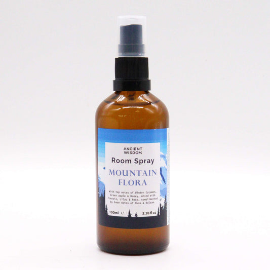 A brown glass bottle of room spray sits on a white background. The label reads: ANCIENT WISDOM Room Spray MOUNTAIN FLORA. It details scent notes of Winter Cyclamen, Green apple & Honey, Freesia, Lilac & Rose, Musk & Balsam. The bottle is 100ml (3.38 fl oz).