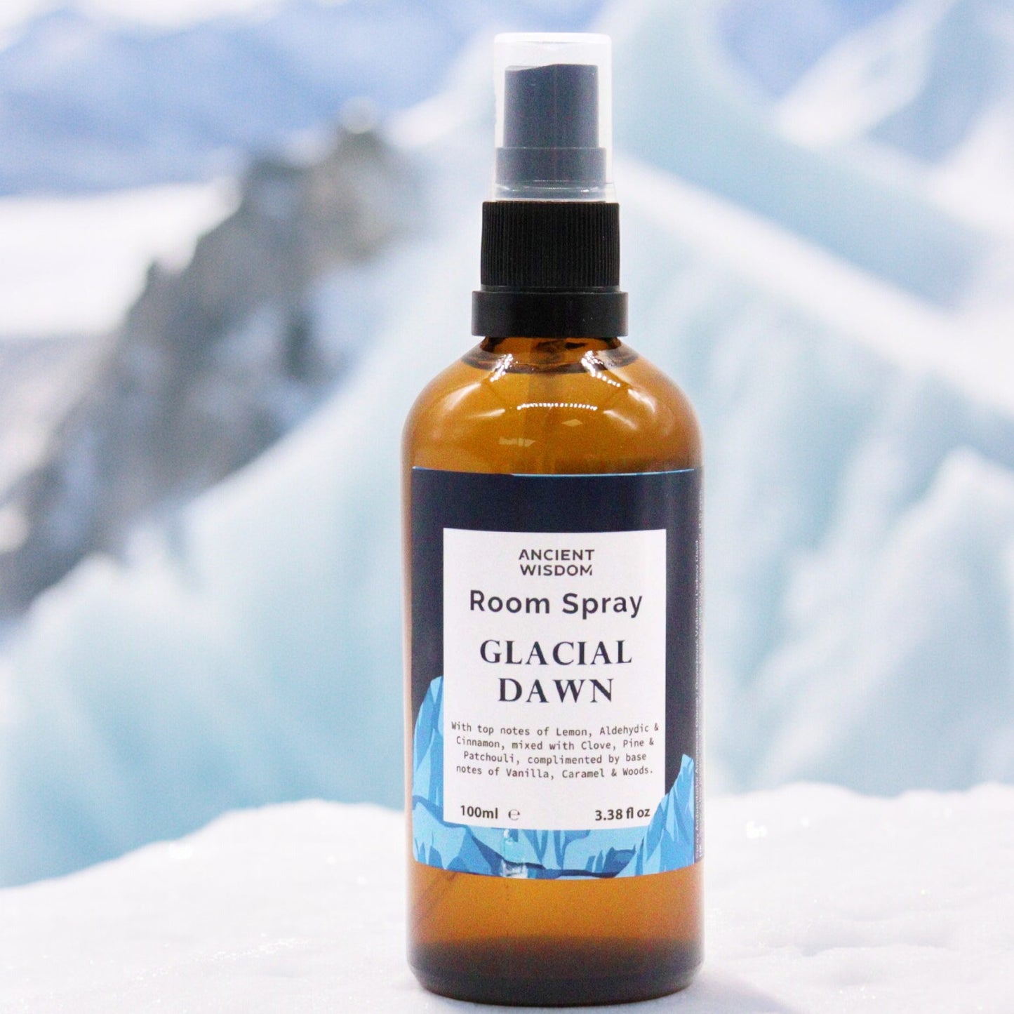 A brown glass bottle of Ancient Wisdom Glacial Dawn room spray sits on a white surface; a blurry icy mountain background is visible.
