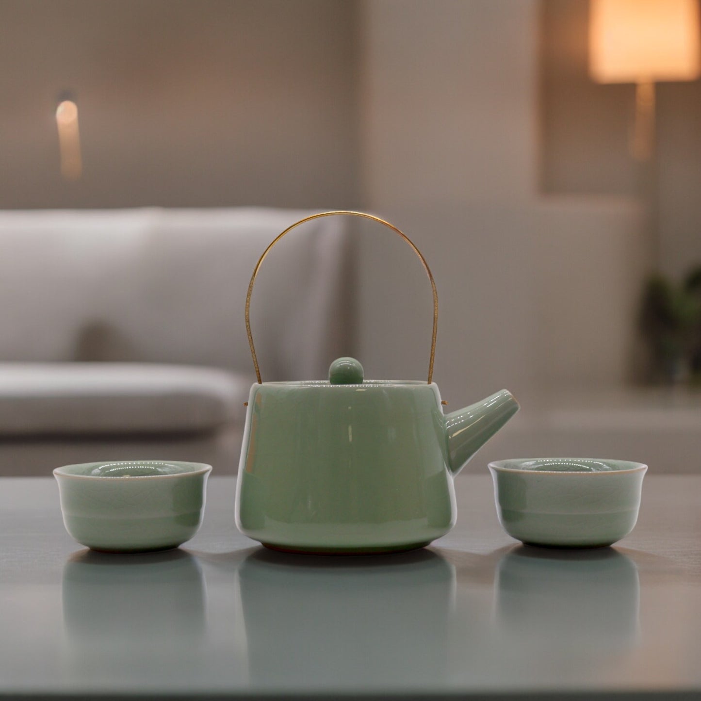 A pale green teapot with a gold handle sits between two matching cups on a reflective table in a softly lit room.