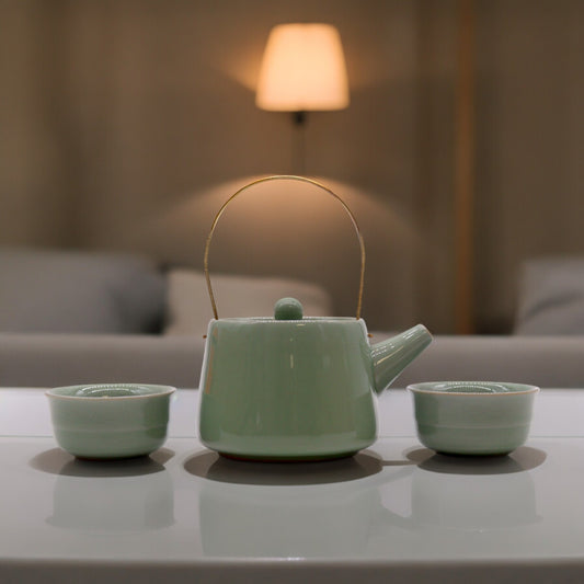 A pale green teapot sits on a white table, flanked by two matching cups; a soft lamp illuminates the scene in a calm, indoor setting.