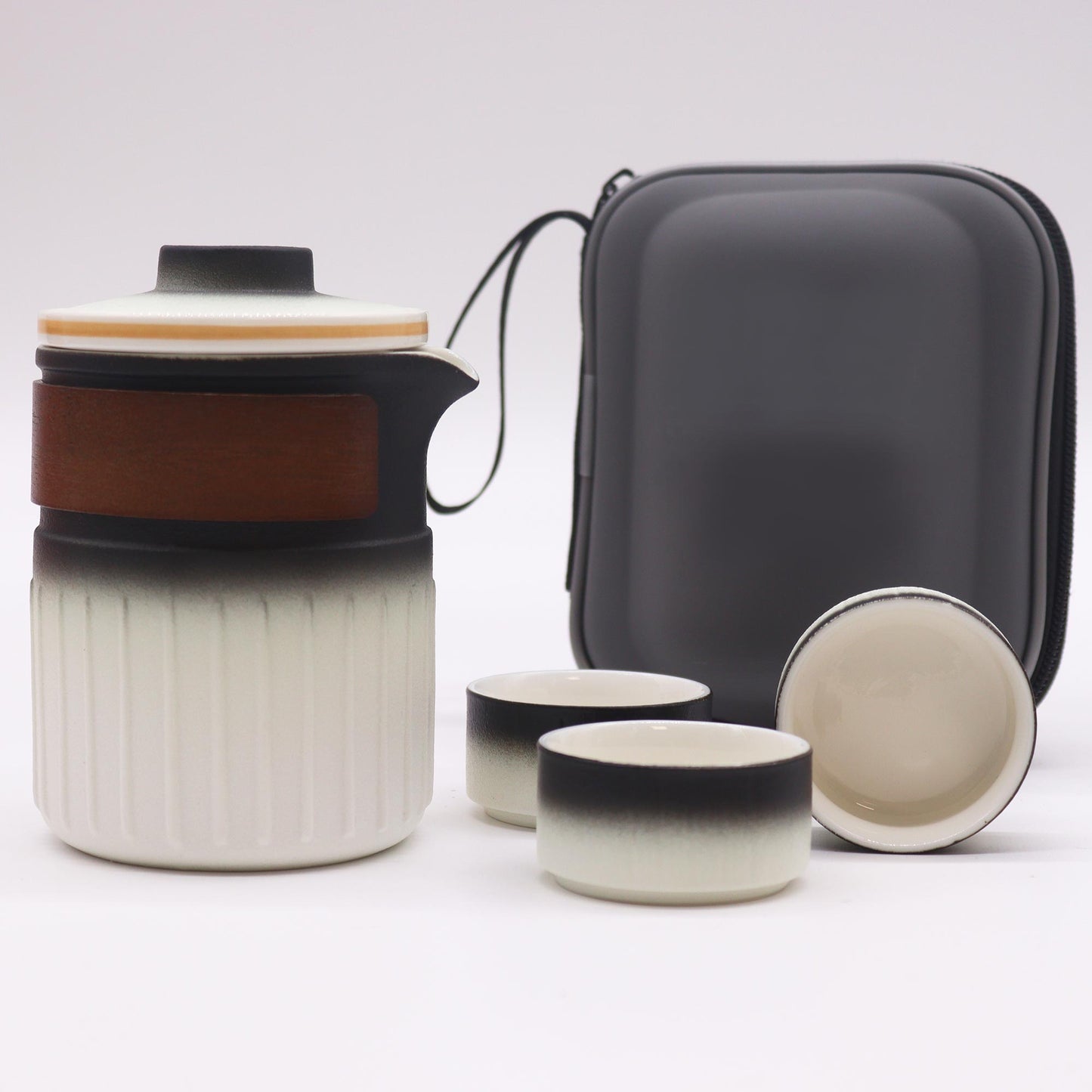 A ceramic tea set, consisting of a teapot and two cups, sits on a white surface next to a gray carrying case. The teapot and cups are predominantly white with black detailing.
