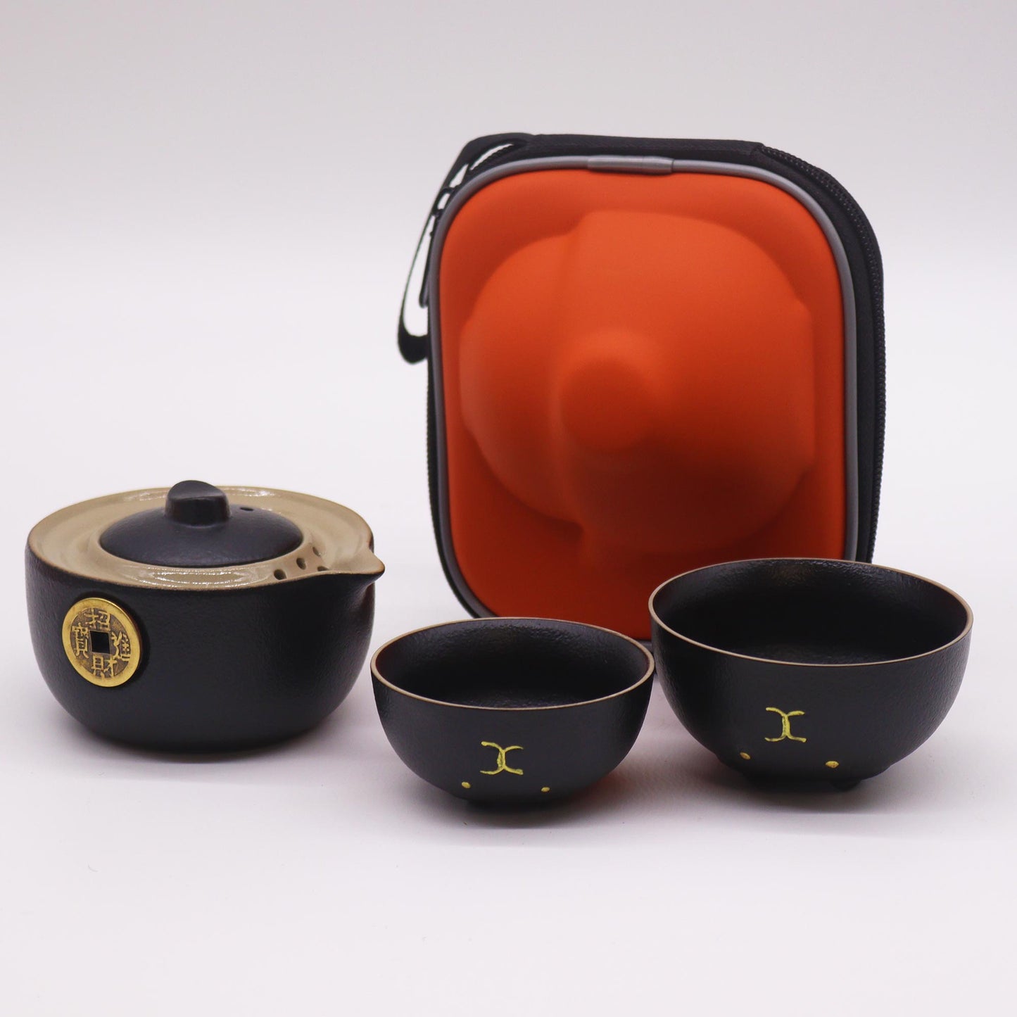 A black ceramic tea set, comprising a teapot and two cups, sits on a white surface alongside an orange carrying case.