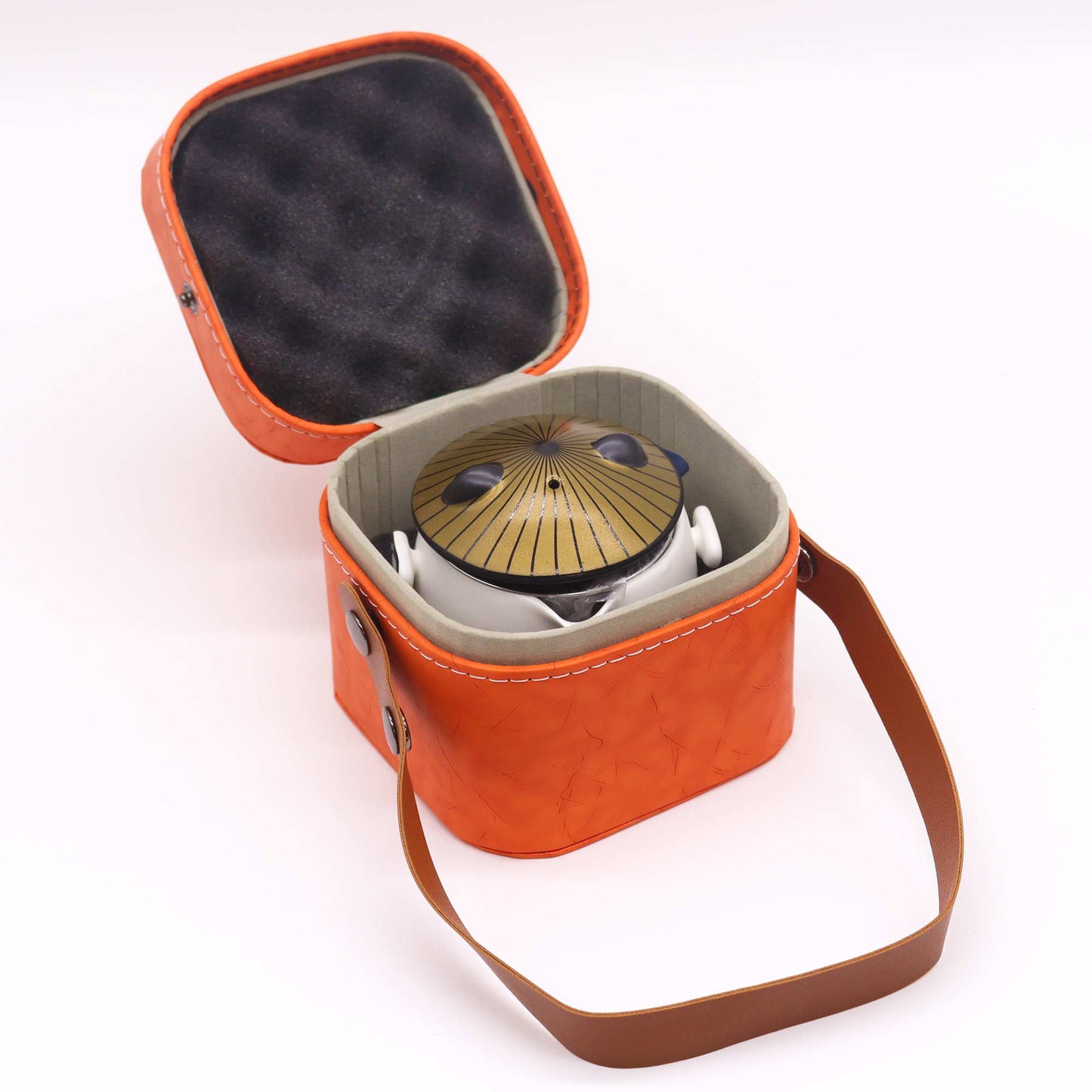 A small, white teapot with a distinctive lid rests inside an orange leather carrying case with a brown strap. The case is open, displaying its contents.