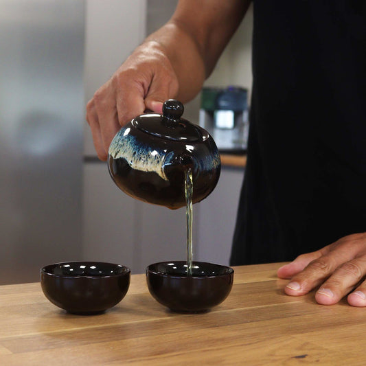 A dark-glazed teapot pours tea into two dark brown bowls on a light wood surface. A person's hands are visible, performing the pouring action in a kitchen setting.