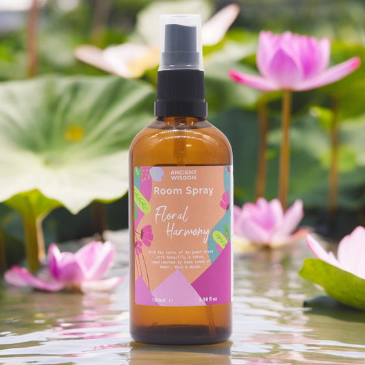 A brown glass bottle containing room spray sits on a surface reflecting pink water lilies; the label reads "ANCIENT WISDOM Room Spray Floral Harmony," detailing fragrance notes.