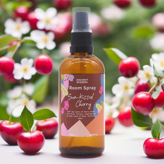 A brown glass bottle of room spray sits among ripe cherries and blossoms. The label reads: "ANCIENT WISDOM Room Spray Sun-Kissed Cherry With top notes of Cherry mixed with Solar Florals, complimented by base notes of Musk, Vanilla & Amber. 100ml e 3.38 fl oz"