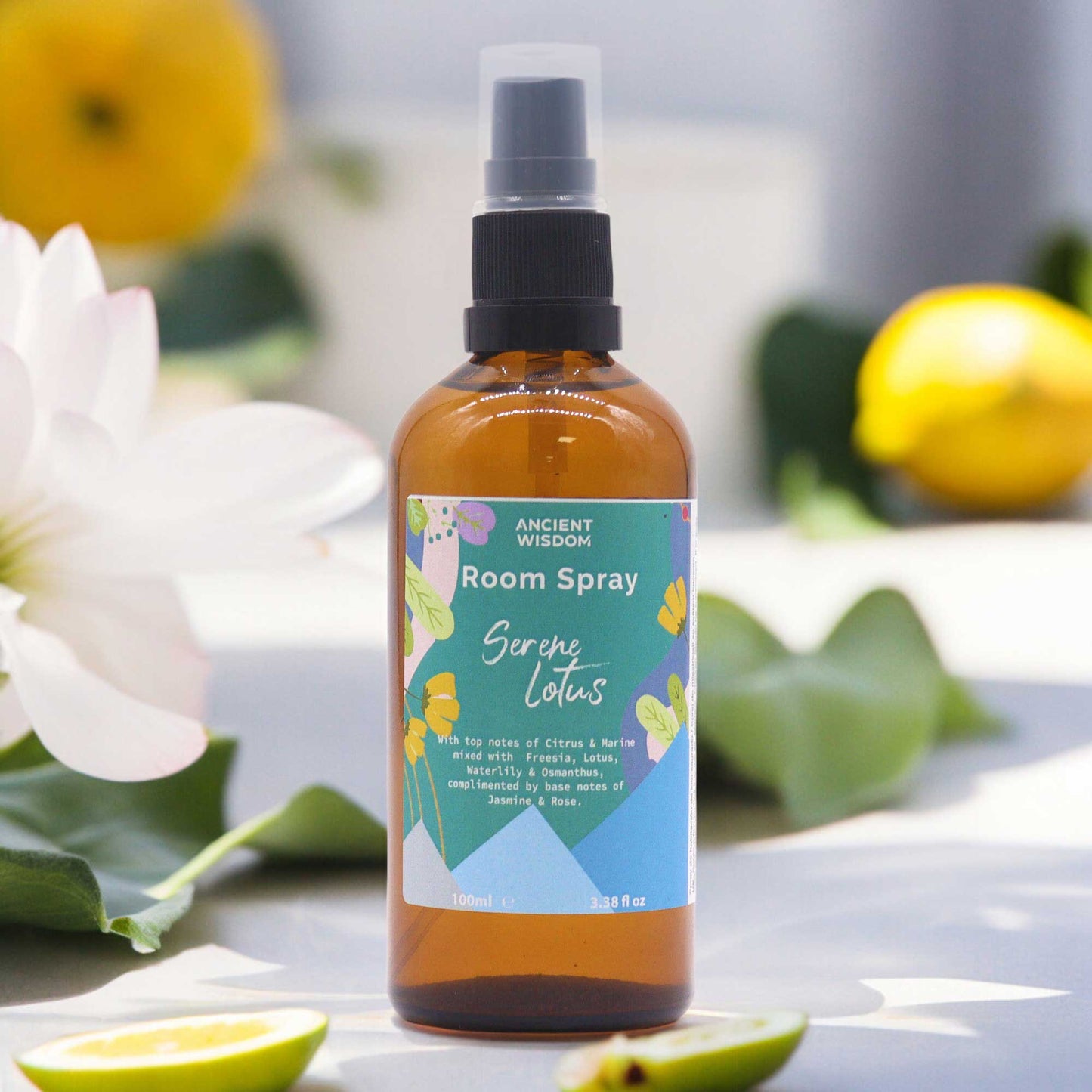 A brown glass bottle of room spray sits on a light surface. The spray bottle is labeled "Serene Lotus," describing its scent profile. Lemons and flowers are softly blurred in the background.