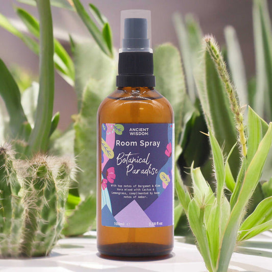 A brown glass bottle of room spray sits among cacti and succulents. The label reads: "ANCIENT WISDOM Room Spray Botanical Paradise With top notes of Bergamot & Aloe Vera mixed with Cactus & Lemongrass, complimented by base notes of Amber. 100ml e 3.38 fl oz".