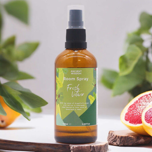 A brown glass bottle of Ancient Wisdom room spray sits on a wooden surface, surrounded by greenery and grapefruit slices. The label reads “Room Spray Fresh Vetiver,” detailing its fragrance notes.