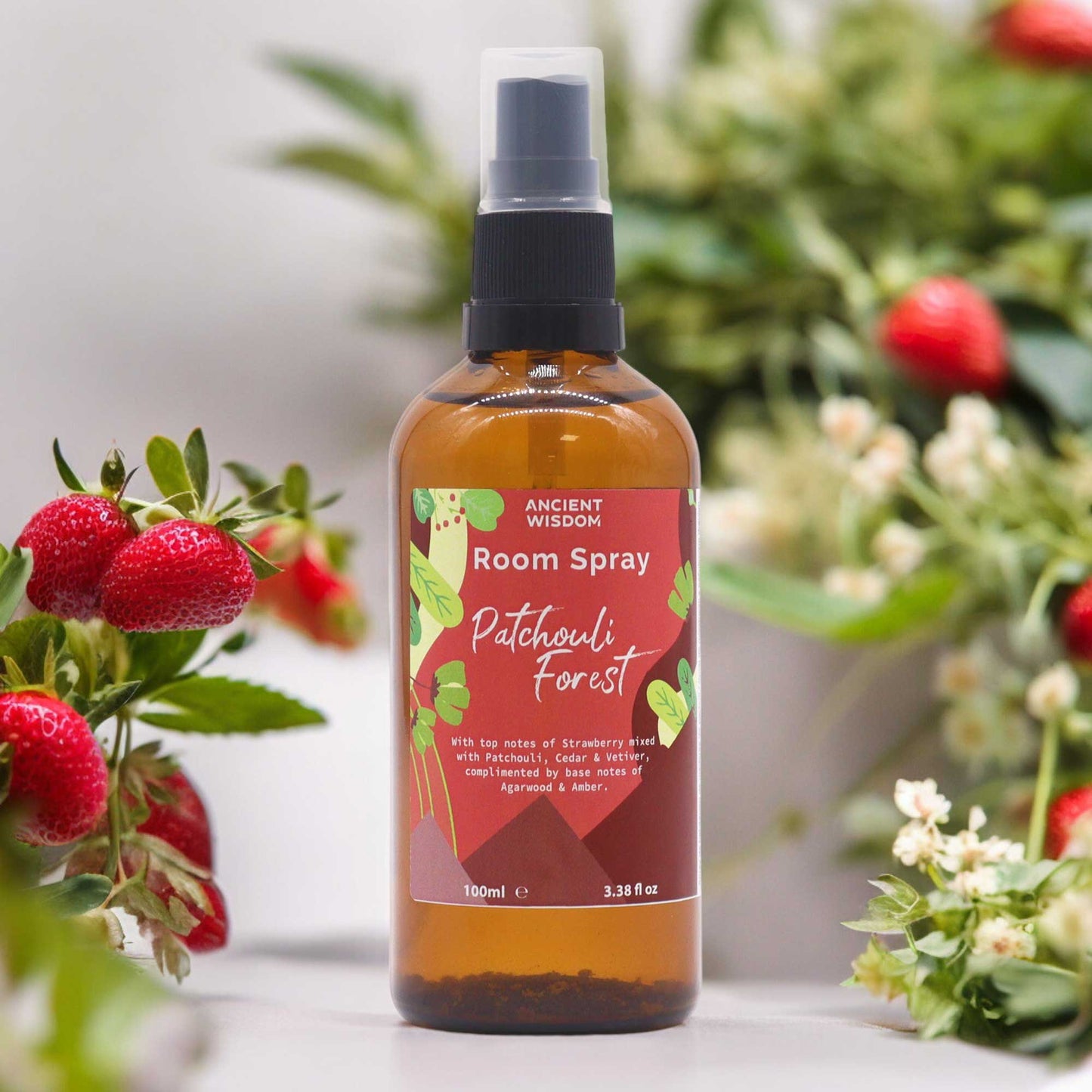 A brown glass bottle of room spray sits amidst strawberries and greenery. The label reads: "ANCIENT WISDOM Room Spray Patchouli Forest With top notes of Strawberry mixed with Patchouli, Cedar & Vetiver, complimented by base notes of Agarwood & Amber. 100ml e 3.38 fl oz".