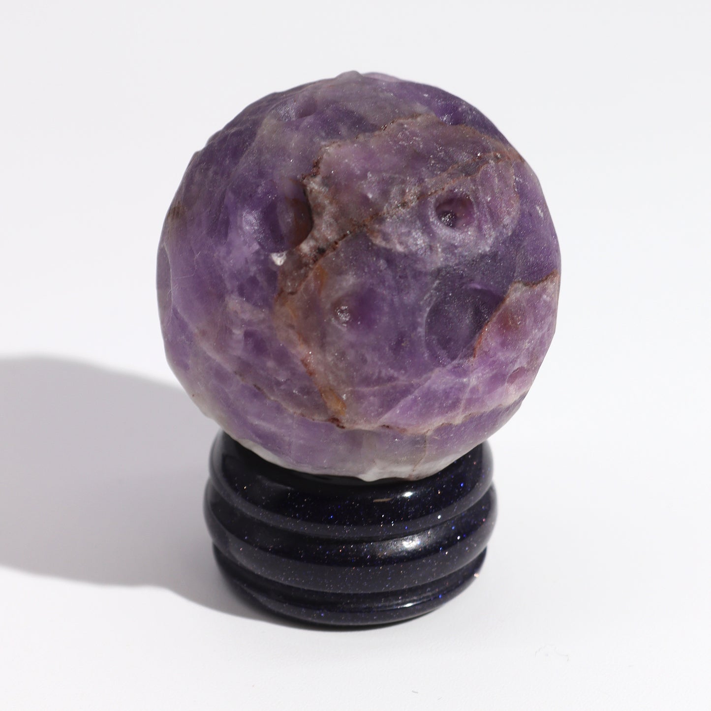 A purple amethyst sphere rests on a dark blue stand. The sphere displays natural texture and variation in color. The context is a plain white background.