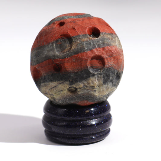 A carved stone sphere, resembling a planet, rests on a dark blue stand. The sphere displays red and black bands and several small holes.