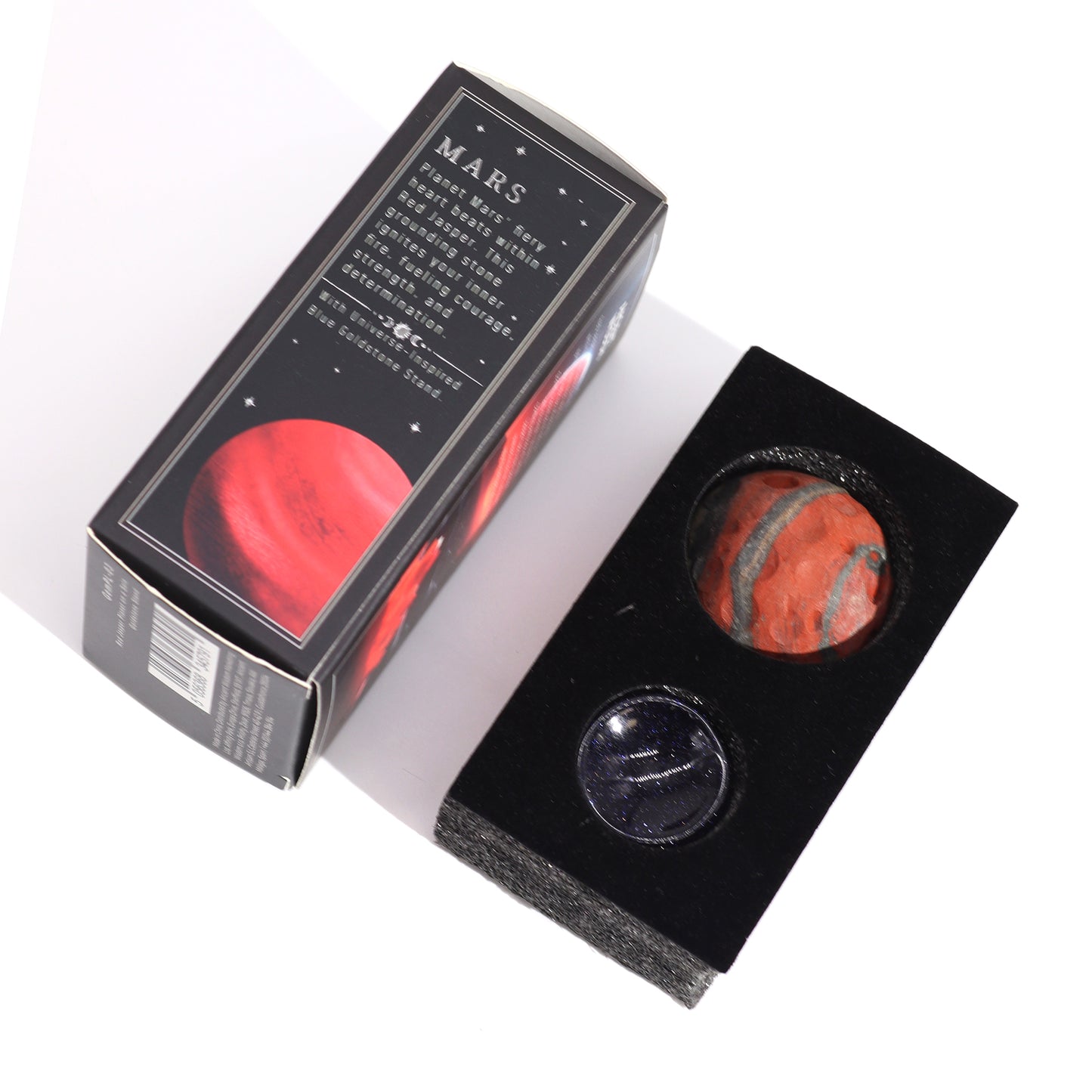Two small spheres, one red jasper and one blue goldstone, rest in a black display case. The case is next to its box, which depicts Mars and details the stones' properties.