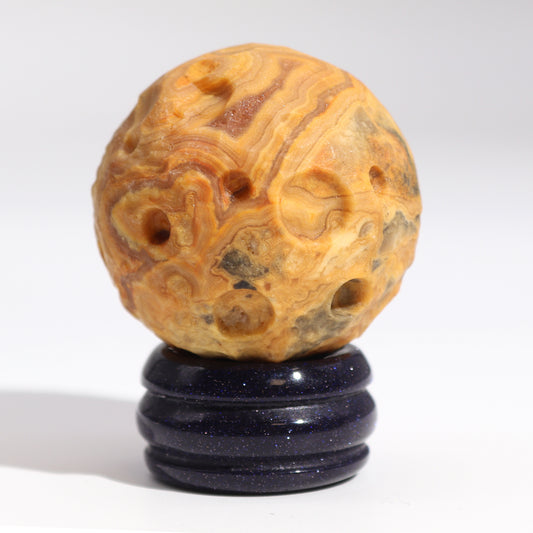 A spherical, orange and brown stone, possibly agate, rests on a dark blue stand. The stone has numerous small holes. It sits on a white background.