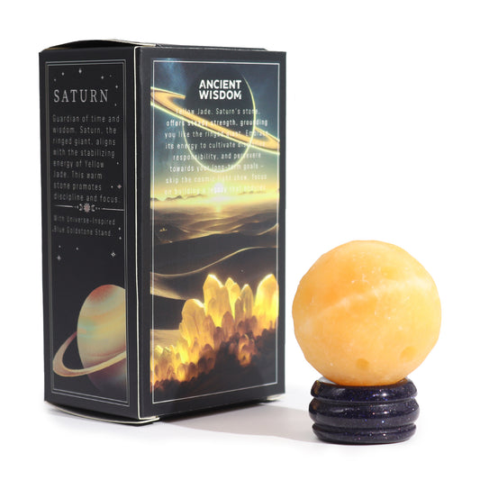 A yellow jade sphere rests on a blue goldstone stand; it sits beside its packaging, which displays astrological imagery and text about Saturn and discipline.