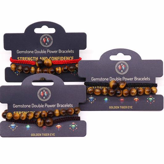 Three gemstone bracelets, displayed on cards, are shown. Each bracelet features golden tiger eye beads and a cord; one cord is red, and the others are dark brown and black. The cards advertise "Gemstone Double Power Bracelets," with one also specifying "Strength and Confidence" and "Golden Tiger Eye."