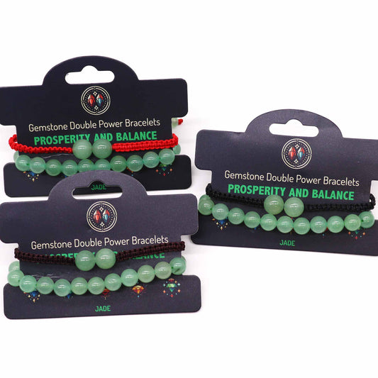 Three jade gemstone bracelets, displayed on individual cards, are presented against a white background. Each bracelet features light green beads and a colored string (red, black, brown). The cards advertise "Gemstone Double Power Bracelets," "PROSPERITY AND BALANCE," and "JADE".