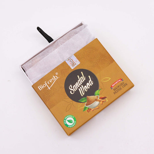 A sandalwood incense cone box sits on a white surface. The box is partially opened, revealing tissue paper inside. The box is gold, and features the text: "Biofresh®," "Sandal Wood," "Charcoal Free," "Backflow Incense Cone," and "Eco Friendly Product."