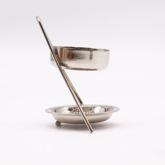A metallic incense burner, consisting of a small cup suspended above a dish by a single, angled rod, sits on a plain white background.