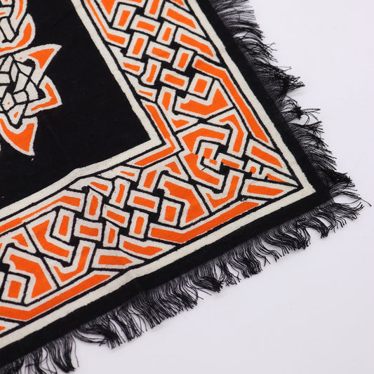 A black textile with orange and white geometric patterns rests on a white surface; its edge shows a black fringe.