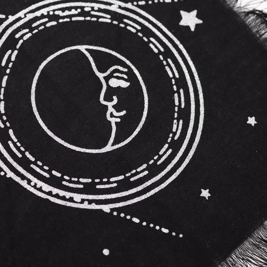 A black textile features a silver-printed crescent moon face within concentric circles and scattered stars; a fringed edge is visible.