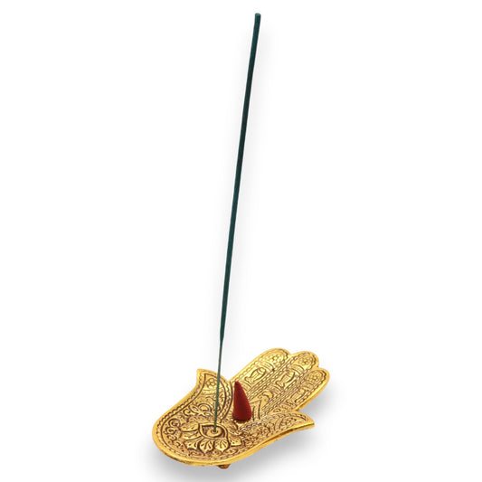 An intricately designed gold-toned hand-shaped incense holder supports a thin, vertical incense stick and a lit incense cone, set against a plain white background.