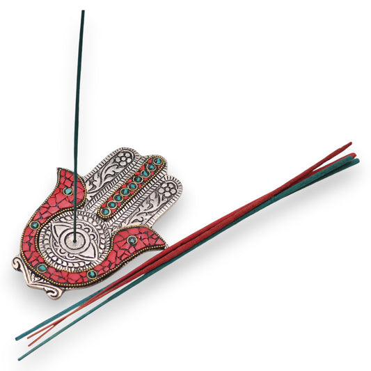 An ornate incense holder shaped like a hand holds a lit stick, with several unlit incense sticks beside it. The hand is decorated with intricate patterns and turquoise accents.