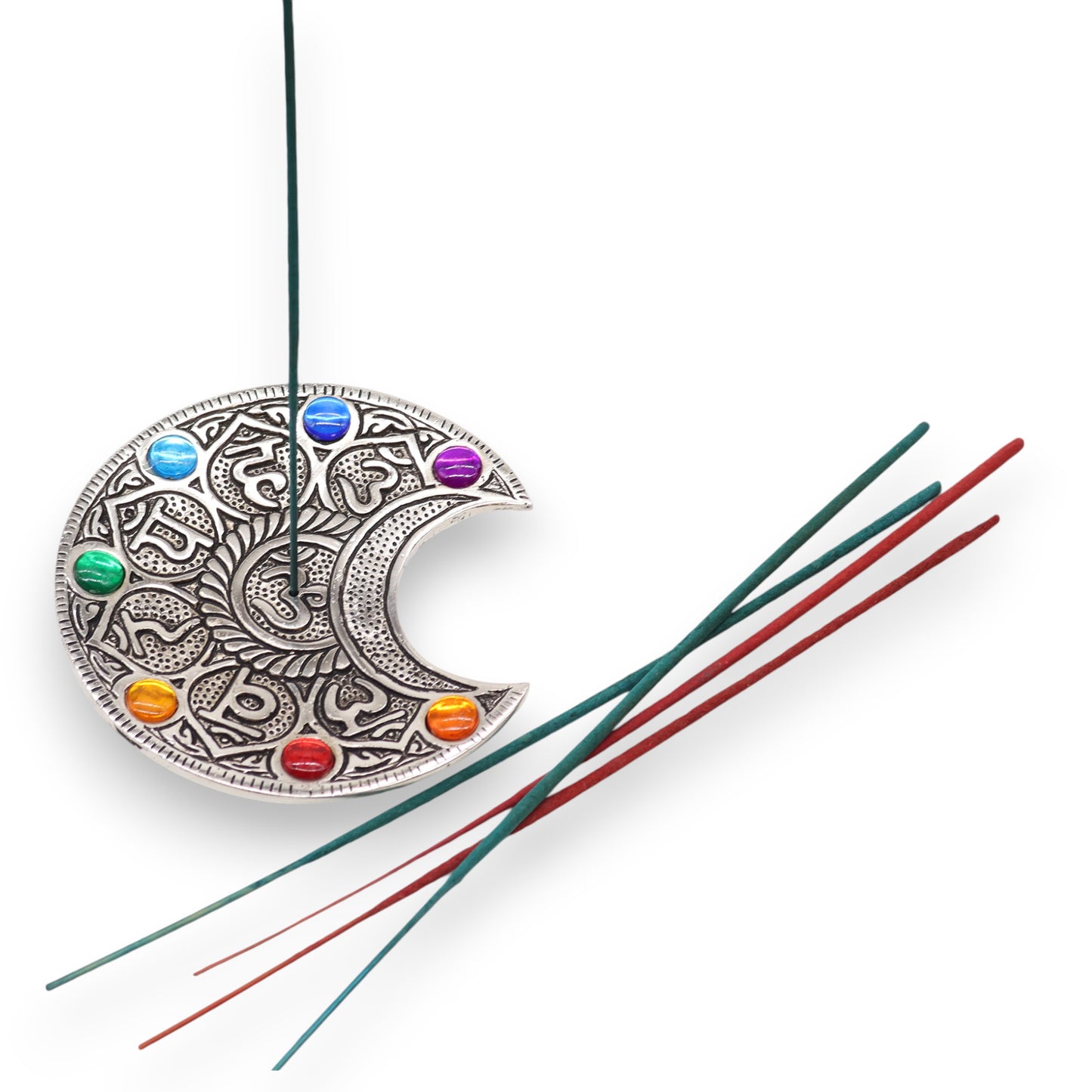 An ornate crescent-shaped incense holder with colorful gems holds a burning green incense stick. Several unlit red and green incense sticks lie beside it on a white background.