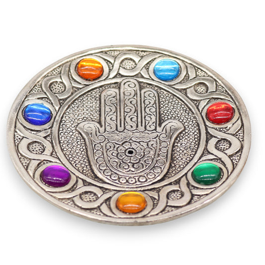 A round, silver metal medallion features an intricately engraved Hamsa hand in the center. It's surrounded by colorful glass beads, set against a background of swirling, engraved patterns.
