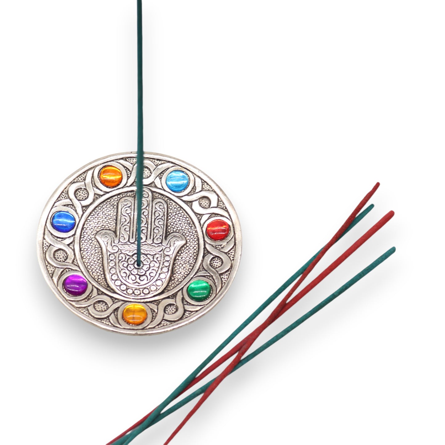 A decorative round incense holder displays a hamsa symbol surrounded by colorful gems, with incense sticks inserted and resting nearby. The white background emphasizes the ornate design.