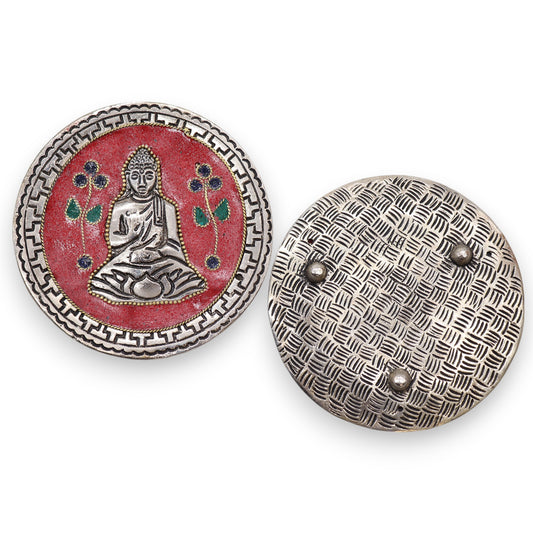 Two round silver shield brooches, one featuring a Buddha figure sitting with floral motifs on a red background, the other displaying a crisscross pattern with three decorative studs.
