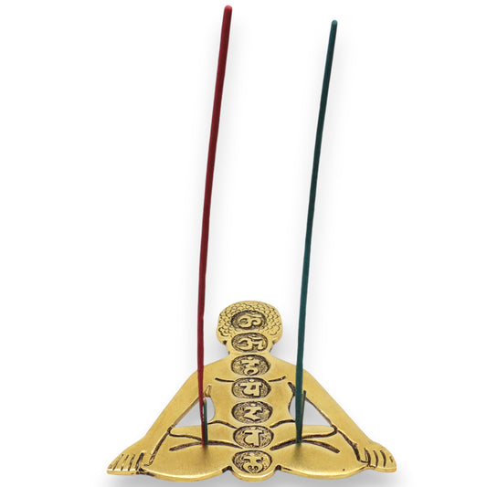 A golden incense holder shaped like a meditating figure holds two lit incense sticks. Symbols are engraved on the figure's body, representing chakras. The background is plain white.