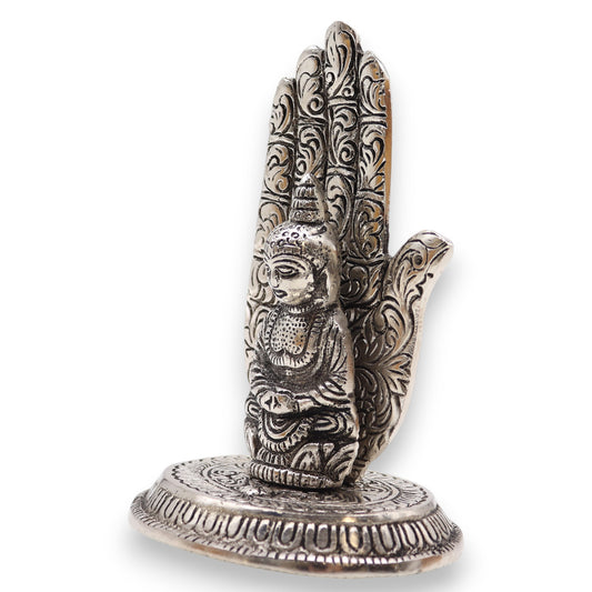 A silver ornamental hand upright on an intricate base, intricately engraved with floral patterns and featuring a Buddha figure in meditation at the palm's center, against a plain background.