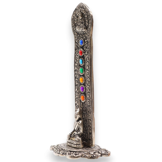 A silver incense holder features intricate engravings and colored stones in a vertical line. A small, seated Buddha figurine is at the base, on an ornate circular platform.