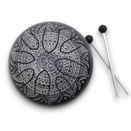 A round, black steel tongue drum features intricate, silver mandala patterns and comes with two mallets placed nearby on a white background.