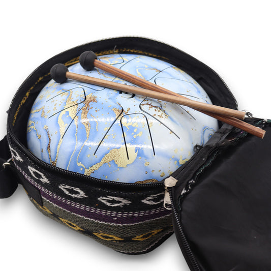 A blue steel tongue drum, with gold flecks and black slits, rests inside an open zippered bag with patterned fabric. Two wooden mallets lie on top of the drum.