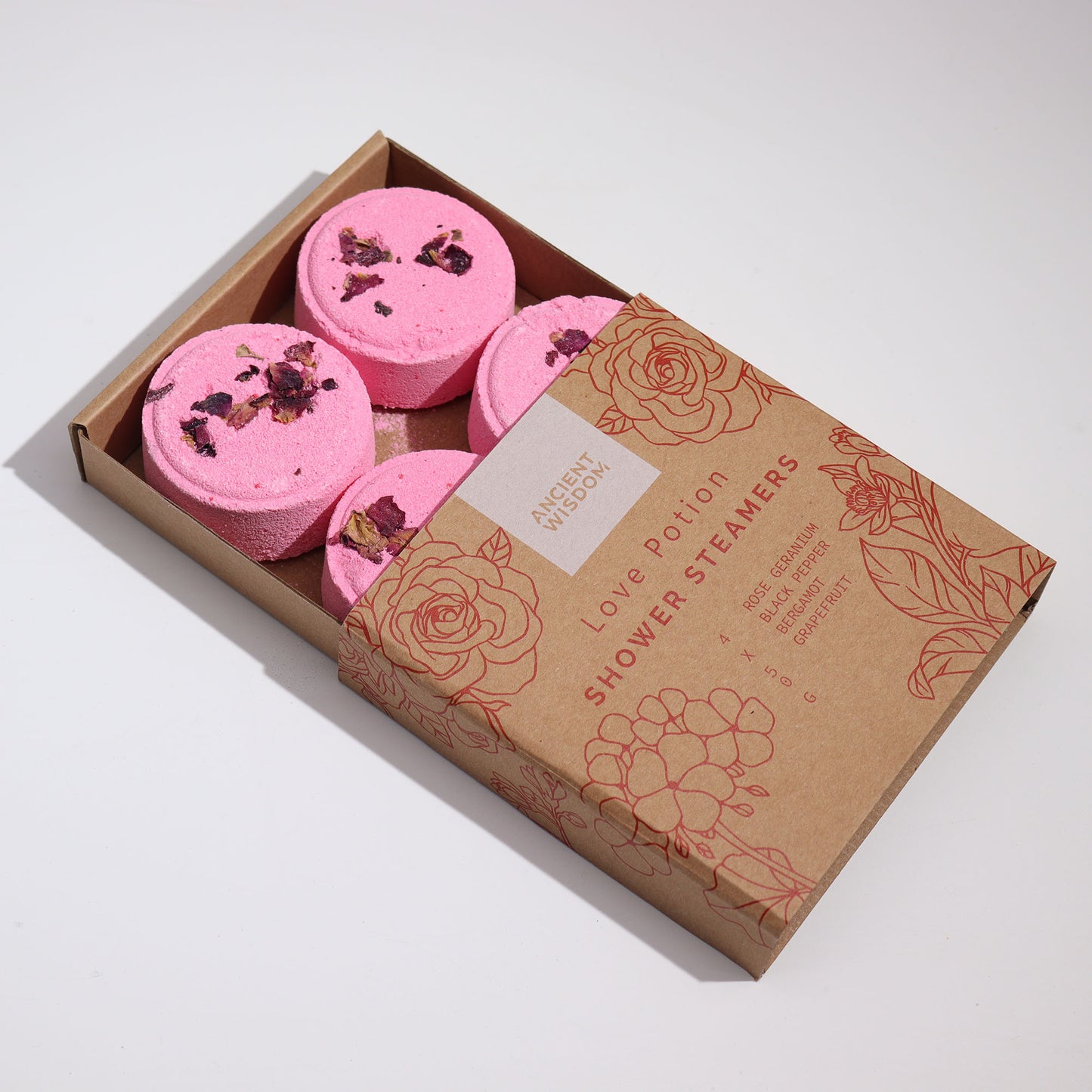 Box with four pink shower steamers topped with dried petals, labeled "Ancient Wisdom Love Potion Shower Steamers," including ingredients: rose geranium, black pepper, and grapefruit. Brown box features floral illustrations.
