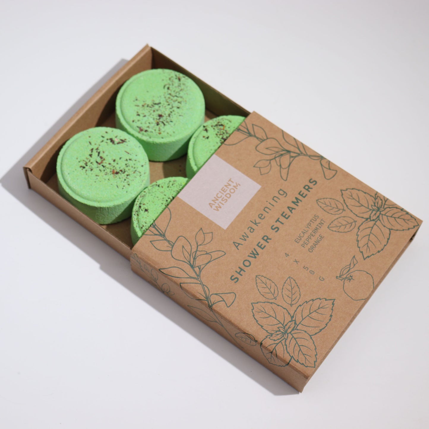 Green shower steamers rest in an open, brown cardboard box. Text on the box reads: "ANCIENT WISDOM, Awakening SHOWER STEAMERS, EUCALYPTUS, PEPPERMINT, ORANGE." Leaf illustrations decorate the packaging.