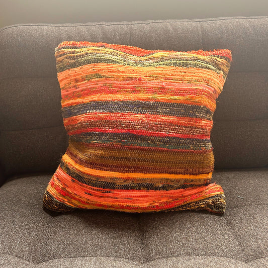 A colorful, textured pillow with orange and red horizontal stripes rests on a dark, cushioned sofa, catching ambient light in a cozy indoor setting.