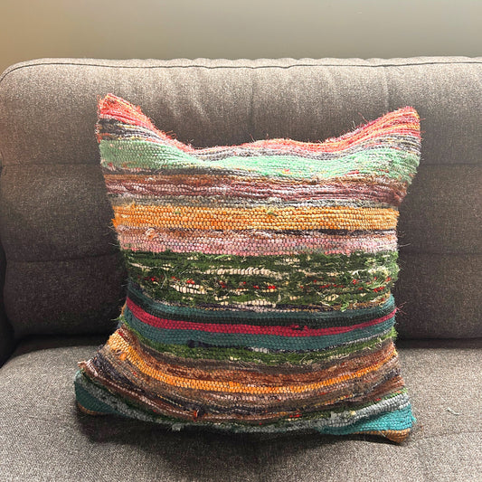 Multicolored woven pillow sits on a gray sofa, featuring horizontal stripes in various shades, including green, pink, yellow, and red, providing a vibrant pattern against the neutral backdrop.