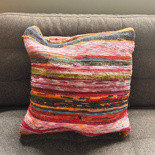 A colorful woven pillow rests against a dark gray sofa, featuring horizontal stripes in pink, red, green, and orange hues, creating a vibrant, textured appearance.