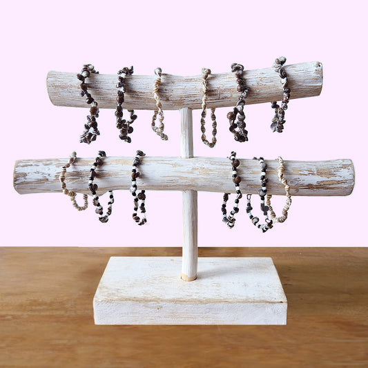 Wooden bracelet stand displaying several twisted rope and bead bracelets. The stand features two horizontal bars and rests on a square base, set against a light pink background.