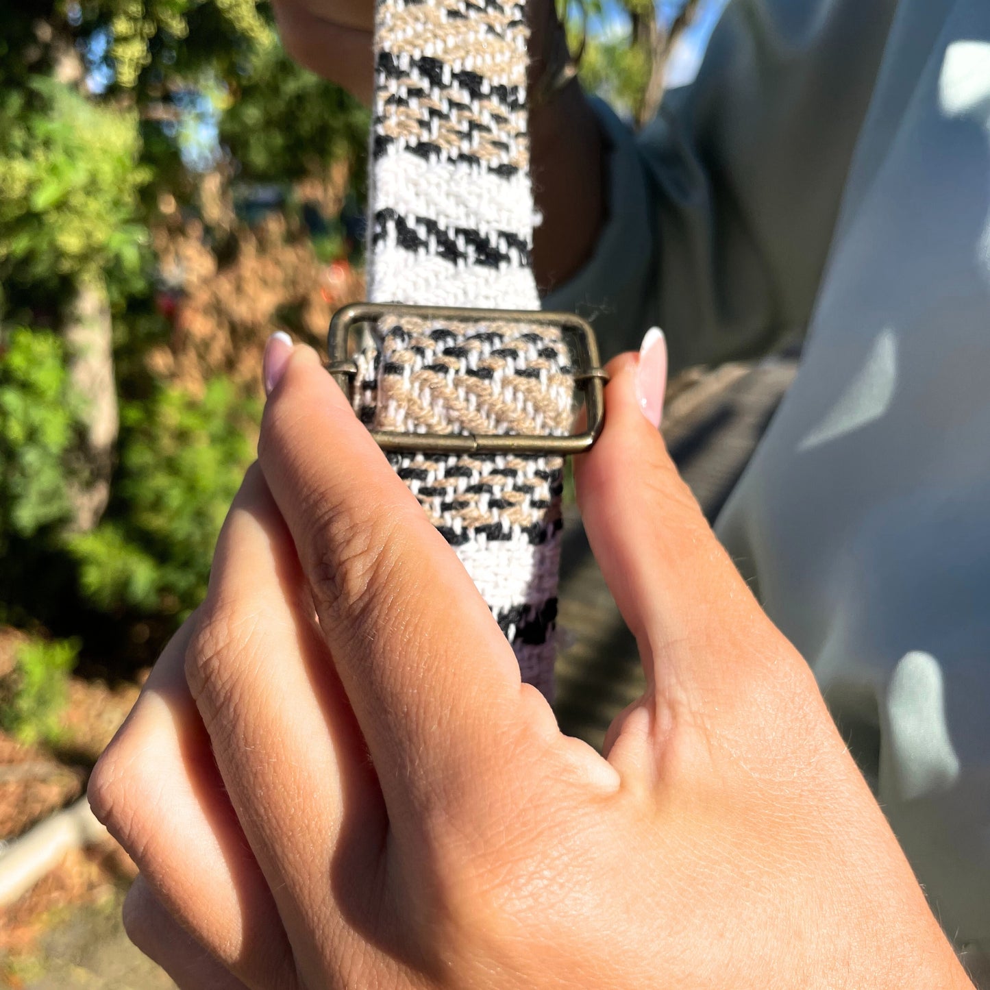 Hands adjusting a woven strap through a metal buckle. The strap has a black, white, and beige pattern. The context is an outdoor setting with greenery and sunlight.
