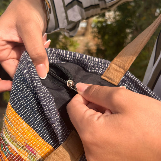 Hands zip closed a colorful fabric bag in a sunny outdoor setting, with green foliage visible in the background.